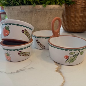 May include: A set of white ceramic bowls with terracotta rims and bases. Two small round bowls are stacked, while a larger dual-compartment bowl features a central terracotta handle. The exterior is decorated with painted tomatoes, asparagus, and peas.