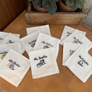 May include: A set of white square fabric napkins featuring embroidered text and illustrations representing different decades, including The Twenties, The Thirties, The Fifties, The Sixties, The Seventies, The Nineties, 2000s, and the Future.