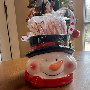 May include: A snowman-shaped ceramic container filled with candy canes. The container has a black top hat, a red scarf, and rosy cheeks. A white ribbon with red polka dots is attached to the container.
