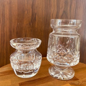 May include: Two clear crystal glass vases with diamond-cut patterns. One vase is short and squat, while the other is taller and rests on a pedestal base. The vases are displayed on a wooden surface.