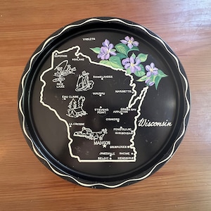 May include: A black serving tray with a white outline of Wisconsin, featuring city names and illustrations. The tray has a scalloped edge and is decorated with purple violets. The word "Wisconsin" is written in white.