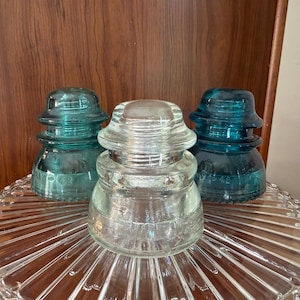 May include: Three glass electrical insulators arranged in a cluster. Two are a deep teal blue color, and one is clear glass. Each insulator features a tiered, rounded shape with a smooth top and a wide base, displaying embossed text on the sides.
