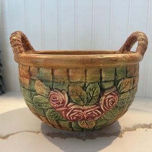 May include: A ceramic bowl with two handles, decorated with a woven basket pattern and pink roses. The bowl is glazed in shades of brown, green, and pink.
