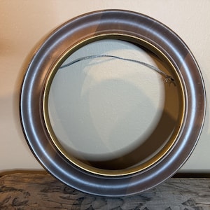 May include: A round picture frame with a metallic silver and bronze finish. The frame has a thick, rounded profile and a gold inner edge. A thin wire is strung across the back for hanging. The frame is resting on a piece of weathered wood.