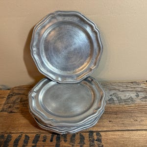 May include: A stack of four round silver-toned metal plates with a smooth, matte finish and a scalloped, wavy edge design. The plates feature a slightly raised rim and a flat center, showcasing a consistent metallic texture throughout the set.