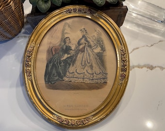 May include: An oval gold-framed picture with a vintage print of two women in 19th-century clothing. The print is titled "La Mode Illustree" and features a woman in a long, white dress with a lace trim and another woman in a dark green dress. The picture is on a white surface.