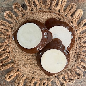 May include: Three brown ceramic ramekins with white interiors. The ramekins are sitting on a woven brown and beige placemat.