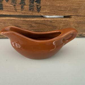 May include: A glossy, brown ceramic gravy boat with a handle and a pouring spout. The boat has a curved design and is set against a wooden background. This kitchenware item is ideal for serving sauces and gravies.
