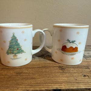 May include: Two white ceramic mugs with a light tan rim. One mug features a green Christmas tree with colorful ornaments, and the other displays a brown Christmas pudding topped with holly. Both mugs are decorated with small gold snowflakes.