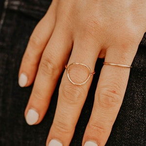 May include: Two gold rings are displayed on a hand. One ring features a delicate open circle design, while the other is a thin band. The hand is resting on a dark denim surface, with a cream-colored sweater in the background.