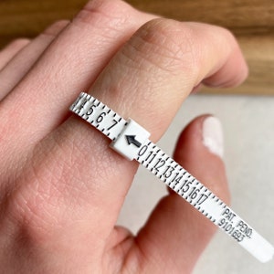 May include: White plastic ring sizer with black numbers from 4 to 17. The sizer is on a finger and the arrow points to the number 11. The text "PAT. PEND. 9101693" is printed on the sizer.