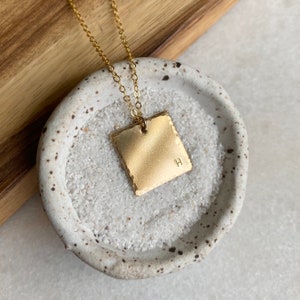 May include: Gold necklace with a square pendant featuring the letter "H" engraved on it. The pendant has a hammered texture and a slightly irregular shape. The necklace is displayed on a white ceramic dish.
