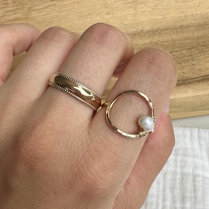 May include: Two gold rings on a finger. The ring on the index finger is a simple band. The ring on the middle finger is a circle with a pearl.