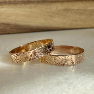 May include: Two gold-colored rings with floral designs. The rings are stacked on top of each other.