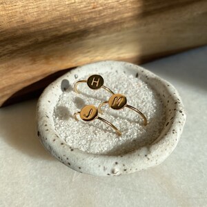 May include: Three gold rings with initial letters H, M, and J on a white ceramic dish filled with white sand.