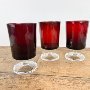 May include: Three ruby red stemmed glasses with a rounded bowl and a clear, circular base. The glasses are arranged on a wooden surface, with a white background. The glasses have a gradient effect, with a darker shade at the bottom.