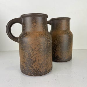 May include: Two brown ceramic jugs with handles. The jugs have a speckled, textured surface with a dark brown colour. The handles are rounded and attached to the top of the jug. The jugs are cylindrical in shape and sit on a white surface.