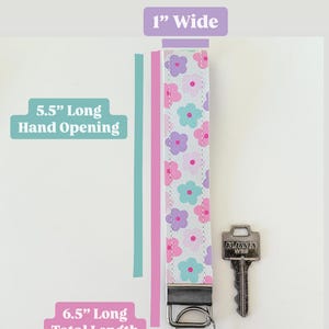 May include: A pink and white key wristlet with a floral pattern. The wristlet is 1 inch wide, 5.5 inches long for the hand opening, and 6.5 inches long total. The wristlet has a silver key ring and a silver key.