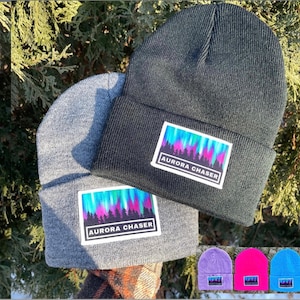 May include: Three knitted beanies with a colourful aurora borealis design and the text "Aurora Chaser". The beanies are grey, black and purple.