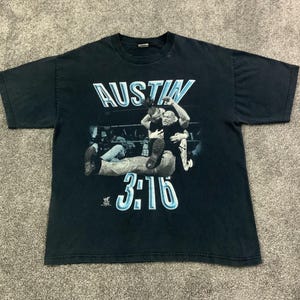 Puede incluir: Camiseta negra con el texto "AUSTIN" en blanco y azul, y un gráfico de dos luchadores. El número "3:16" está impreso debajo del gráfico en azul. La camiseta está hecha de un material suave.