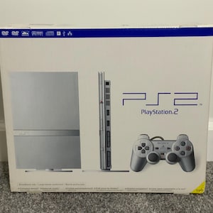 May include: A white PlayStation 2 game console box featuring a silver console shown from the front and side, along with a silver controller. The box displays the PS2 logo in blue text and includes various technical format icons at the top edge.