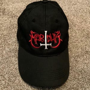 Puede incluir: Gorra de béisbol negra con la palabra "Marduk" bordada en rojo, con letras estilizadas y una cruz blanca. La gorra tiene una visera curva y un botón en la parte superior. El sombrero es de tela oscura.