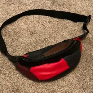 May include: A black fanny pack with a red, brown, and black patchwork design. The bag has a zippered closure and an adjustable strap.
