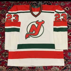 Puede incluir: Una camiseta de hockey blanca con detalles rojos y verdes. La camiseta presenta el número 23 en rojo en las mangas y un logotipo del equipo en el pecho. Tiene cuello en V y probablemente está hecha de un material transpirable.