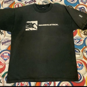 May include: Black t-shirt featuring the band name "MASSIVE ATTACK" in white text. The shirt has a graphic design on the front, and a small tag on the sleeve. The shirt is displayed on a baseball-themed background.