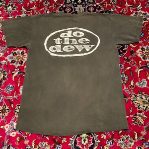 Pode incluir: Uma camiseta cinza escuro com a frase "do the dew" em branco dentro de um oval desgastado. A camiseta está estendida, mostrando a parte de trás. O texto está ligeiramente desbotado, dando um aspecto vintage. A camiseta é de algodão.