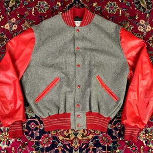 May include: A gray wool varsity jacket with bright red leather sleeves. The jacket features red accents on the collar, pockets, and cuffs, with red button closures. A classic, sporty style.