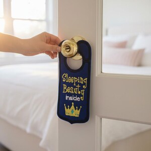 Könnte beinhalten: Ein dunkelblauer Türhänger aus Filz mit schwarzem Rand, der den gelben gestickten Text "Sleeping Beauty inside" und eine gelbe Kronengrafik mit hellblauem Mittelakzent zeigt, hängend an einem runden Türknauf aus poliertem Messing.