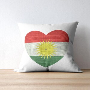 May include: White square pillow with a heart-shaped design. The heart features the colors of the Kurdish flag: red, white, and green, with a yellow sun emblem in the center. The pillow is on a wooden surface.