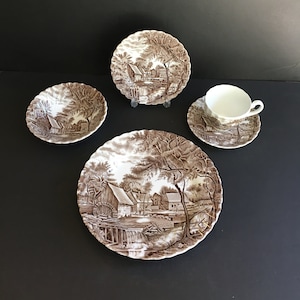 May include: A set of brown and white ceramic dinnerware featuring a country scene with a watermill and trees. The set includes a dinner plate, a salad plate, a bowl, a saucer and a teacup.