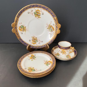 May include: A vintage porcelain tea set featuring a white base, gold trim, and floral patterns. The set comprises a large serving plate, smaller plates, a teacup, and a saucer. The plates have a blue border with gold detailing.