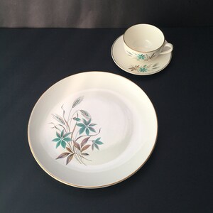 May include: A white ceramic dinner plate with a gold rim and a green and brown floral design. A matching teacup and saucer with the same floral design are also pictured.