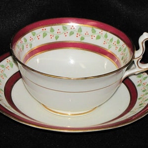 May include: A white teacup and saucer set with a burgundy and gold rim and a green vine pattern.