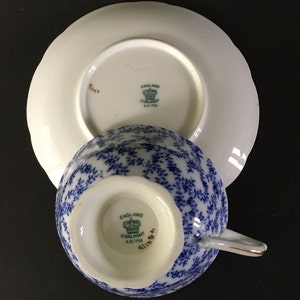 May include: A blue and white teacup and saucer set with a floral pattern. The bottom of the cup and saucer have a green and gold logo that reads "England Coalport A.D.1750".