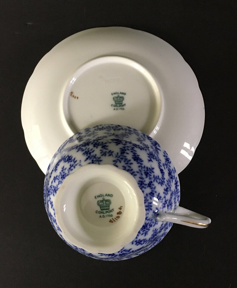 May include: A blue and white teacup and saucer set with a floral pattern. The bottom of the cup and saucer have a green and gold logo that reads "England Coalport A.D.1750".