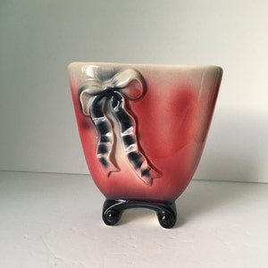 May include: A vintage ceramic vase with a red and cream gradient. A raised bow design with navy blue and cream ribbons adorns the front. The vase has a black base with two curved feet. The vase is likely from the mid-20th century.