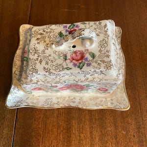Könnte beinhalten: Eine antike Butterdose aus Porzellan mit Blumenmuster und goldener Verzierung. Der Deckel hat einen dekorativen Griff.