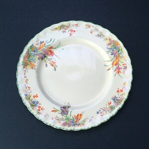 May include: A white ceramic plate with a green rim and a floral design featuring yellow, orange, and purple flowers. The plate has a scalloped edge.