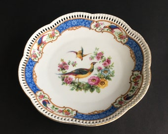 Peut inclure: Une assiette en porcelaine blanche avec un bord bleu et or et une bordure ajourée délicate. Le centre présente un motif floral coloré avec deux oiseaux, un jaune et un bleu, et un faisan plus grand avec un plumage rouge, jaune et bleu.