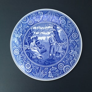 Peut inclure: Une assiette en céramique bleue et blanche avec une bordure florale et une scène de deux personnes dans un jardin. L'assiette est décorée d'un motif floral bleu et blanc.