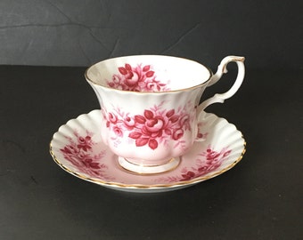 May include: A white teacup and saucer with a pink and red floral pattern. The cup and saucer have a gold trim.