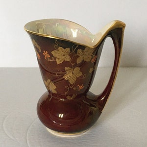 Könnte beinhalten: Eine dunkelrote Keramikkanne mit goldfarbenem und orangefarbenem Blumendesign. Die Kanne hat eine breite Öffnung und einen geschwungenen Griff.