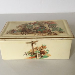 May include: A vintage ceramic box with a cream-coloured base and a lid featuring a colourful illustration of a building with a horse-drawn carriage and people. The lid has a gold trim. The box has a second illustration on the front of a signpost with a tree and bushes.