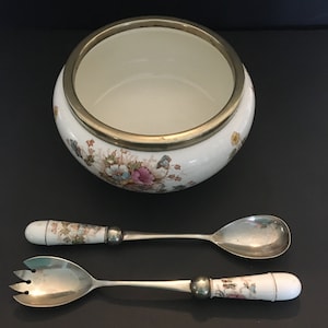 Może przedstawiać: Biała porcelana miska z pozłacanym brzegiem i kwiatowym wzorem, wraz z pasującą srebrną łyżką i widelcem do serwowania z porcelanowymi uchwytami.