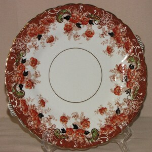 Könnte beinhalten: Ein weißer Keramikteller mit einem gewellten Rand und einem floralen Design in Orange-, Braun- und Grüntönen. Der Teller hat einen goldenen Rand.