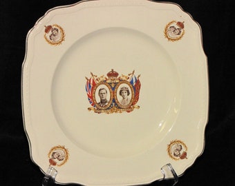 May include: A white square ceramic plate with a gold rim and a royal crest in the center. The crest features a crown, two flags, and portraits of King George VI and Queen Elizabeth. The plate is decorated with four corner designs of a wreath and crown.